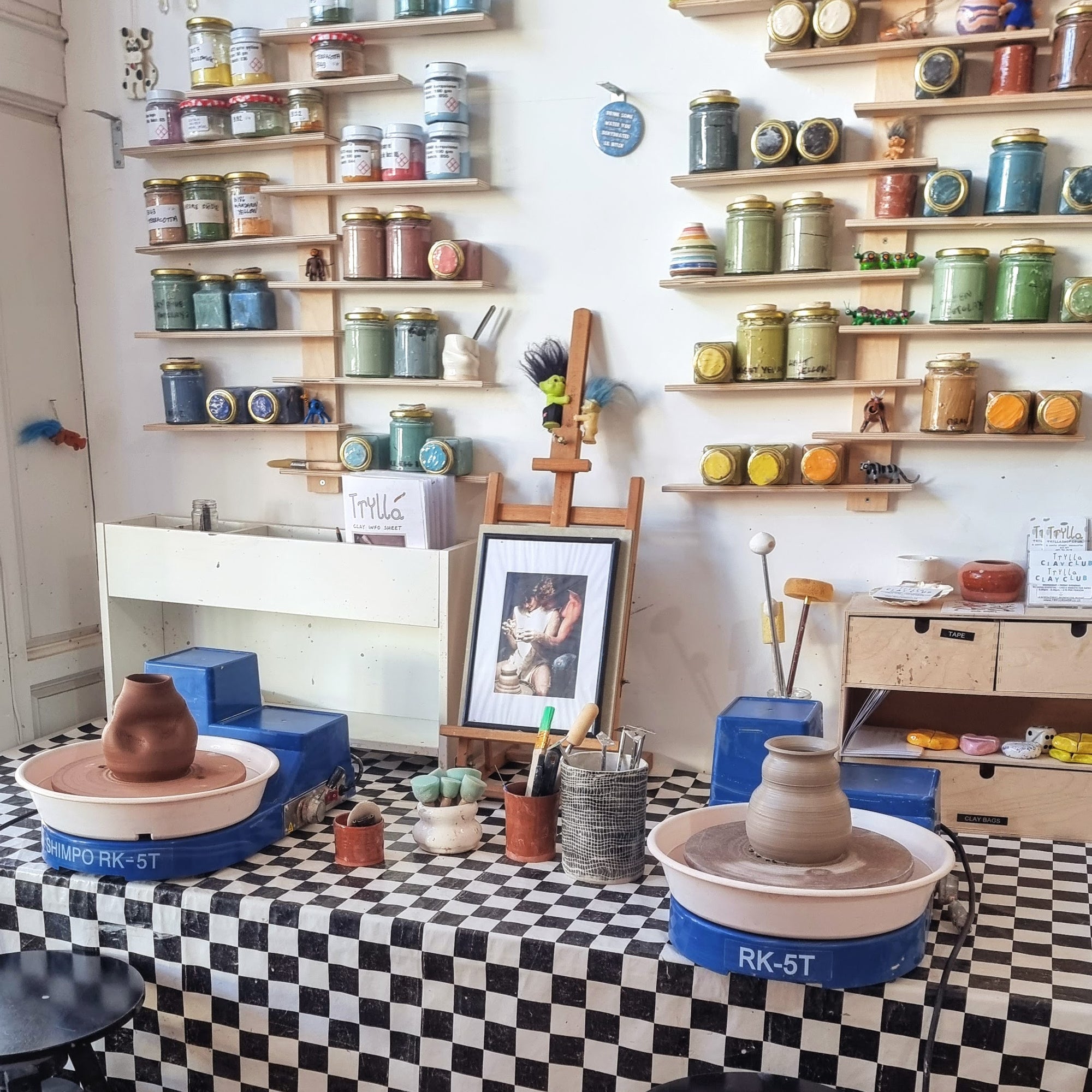 Two pottery wheels with vases set up on a table at Trylla Bristol, with shelves of paints in the background